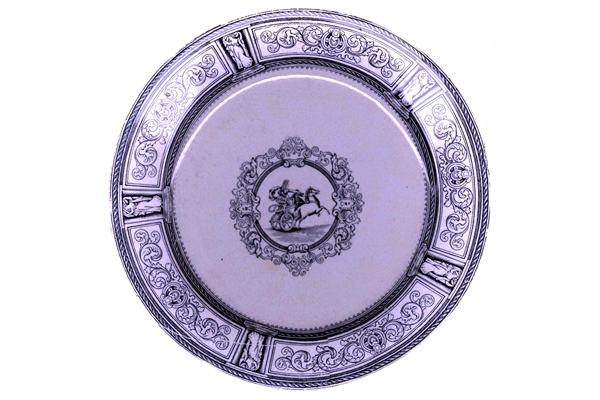 Late 19th century glazed ceramic plate manufactured by the Clyde Pottery Company, Greenock. Decorated with the 'Chariot' pattern of a charioteer, receding, with two prancing horses, in central cartouche, heavily decorated with classical scrollwork; border, scrollwork, narrow reserves with female classical figures. - 1987.597 - © McLean Museum and Art Gallery, Greenock.