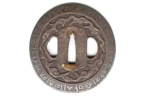 A Japanese swordguard or tsuba, date unknown. The design features a border of archaic letters & symbols inlaid in silver around the rim. The centre has a gilded and carved design of a dragon. - 1981.143 - © McLean Museum and Art Gallery, Greenock