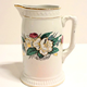 Jug manufactured by the Clyde Pottery Company, Greenock. Decorated with the 'York' pattern. White background with zigzag pattern in relief around rim; narrow gilt band round rim and foot. Band of decoration around middle, floral pattern in green, pink, turquoise and gold. Mark on back 'YORK C.P.Co' inside laurel branches and surmounted by crown. - L1977.142 - © McLean Museum and Art Gallery, Greenock.